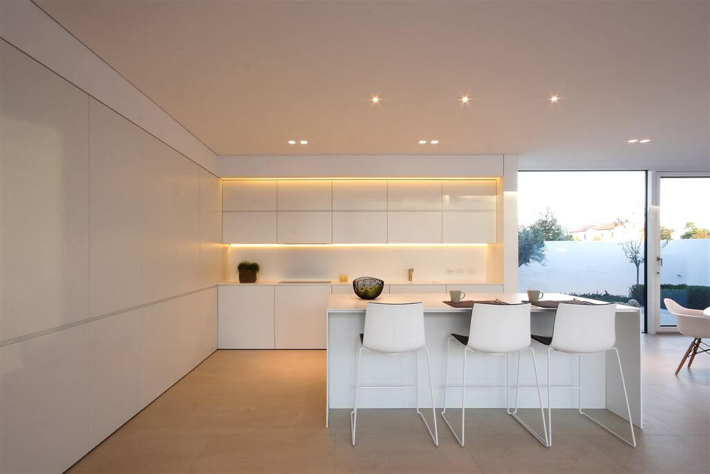 Kitchen area features hidden lighting around cupboards, all-white minimal surfaces, and bar-style seating at island. Kitchen area features hidden lighting around cupboards, all-white minimal surfaces, and bar-style seating at island.