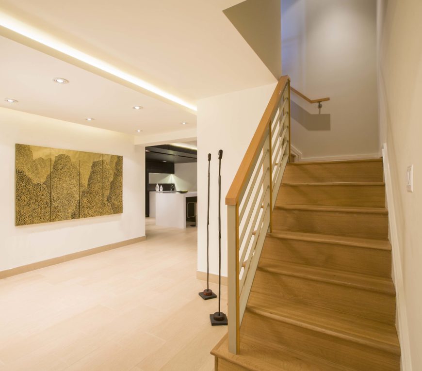 Staircase from main floor, opening to wide hallway with light hardwood flooring, embedded lighting in the drop ceiling, and large art piece on wall. Staircase from main floor, opening to wide hallway with light hardwood flooring, embedded lighting in the drop ceiling, and large art piece on wall.