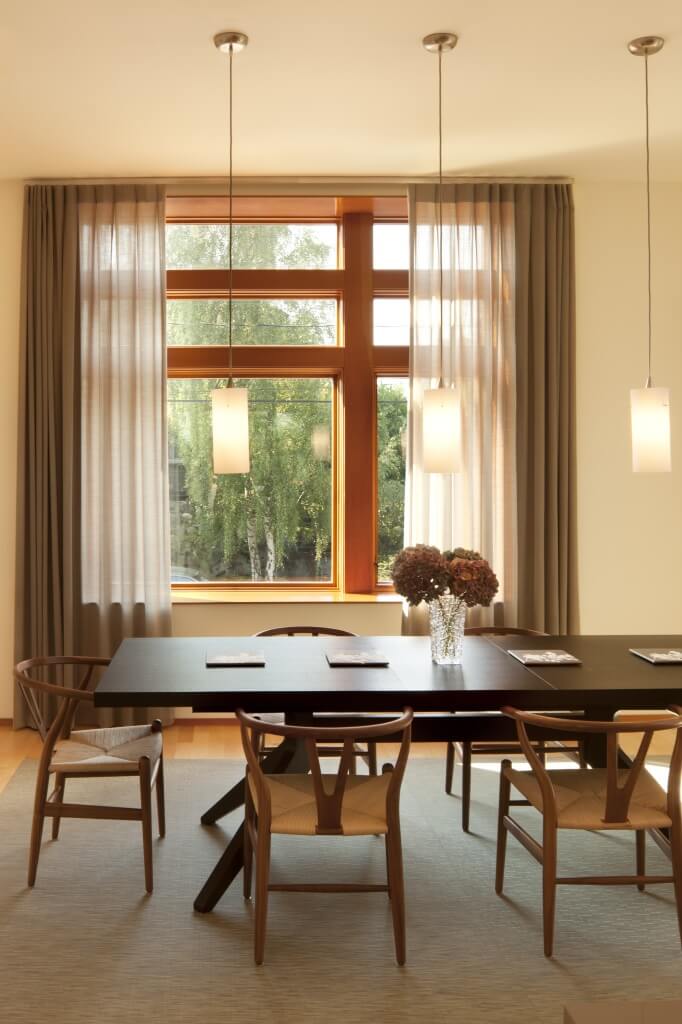 Close view of dining area, with dark wood table pairing with lighter natural wood chairs over a beige rug. Yet another ceiling height window offers natural light, behind hung minimal chandeliers. Close view of dining area, with dark wood table pairing with lighter natural wood chairs over a beige rug. Yet another ceiling height window offers natural light, behind hung minimal chandeliers.