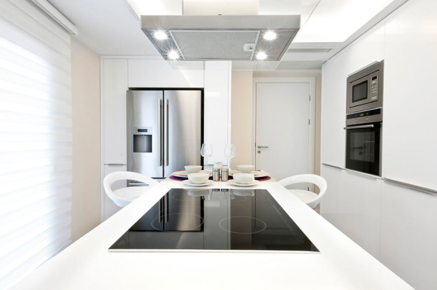 The four small, but bright, spotlights in this stove hood ensure that the cooktop is well lit and the small breakfast bar of the island is lit as well. The impressive amount of white in this kitchen reflects so much from the recessed lighting in the ceiling that the stove hood lights are only necessary for the cooktop.