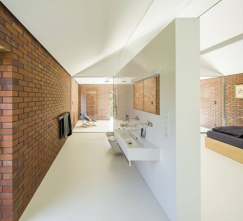 This bathroom takes up a large space, with a large white wall separating the bathroom area from the bedroom. While the walls consist of a traditional red brick, the floors and separating wall are stark white, providing a healthy balance for the space. A trough style floating sink is featured on the inside of the separation wall, as well as a glass walled shower on the far end. This bathroom takes up a large space, with a large white wall separating the bathroom area from the bedroom. While the walls consist of a traditional red brick, the floors and separating wall are stark white, providing a healthy balance for the space. A trough style floating sink is featured on the inside of the separation wall, as well as a glass walled shower on the far end.