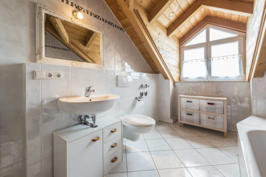 A section of wall protrudes from the surrounding wall, and features a floating sink, a toilet, and a cabinet below the sink for storage. The ceiling contrasts the white tile with its golden wood tones, and the large window lets in plenty of natural light to illuminate the room. A section of wall protrudes from the surrounding wall, and features a floating sink, a toilet, and a cabinet below the sink for storage. The ceiling contrasts the white tile with its golden wood tones, and the large window lets in plenty of natural light to illuminate the room.