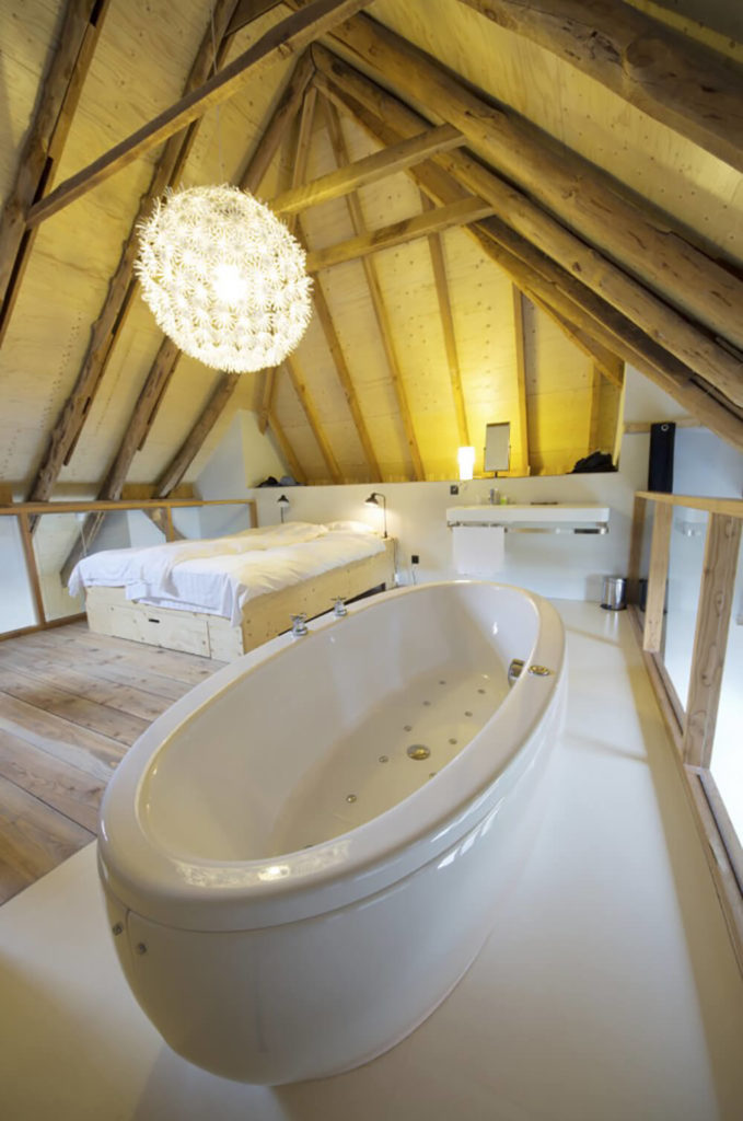 A large luxurious jet tub takes up a good amount of space in this room, and sits upon a light tile floor. You can see from the walls that this space sits on a loft in the upper section of the home. There is a floating vanity on the far end of the room. A large bed sits on a natural wood frame, matching the hardwood floor in the rest of the space. A large luxurious jet tub takes up a good amount of space in this room, and sits upon a light tile floor. You can see from the walls that this space sits on a loft in the upper section of the home. There is a floating vanity on the far end of the room. A large bed sits on a natural wood frame, matching the hardwood floor in the rest of the space.