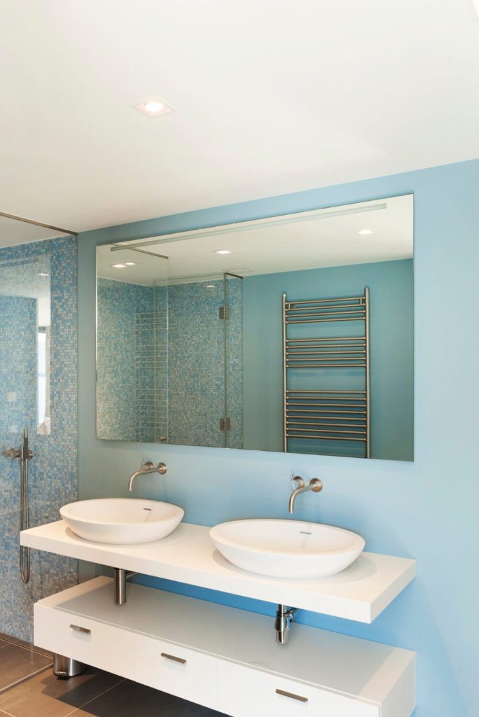 Small square blue tiles cover the interior of the shower, while the walls transition to a solid baby blue behind the sinks and mirror. Beneath the floating sinks there is a set of white drawers that provide plenty of storage space. In the reflection of the mirror, you can see a chrome towel rack on the opposite wall. Small square blue tiles cover the interior of the shower, while the walls transition to a solid baby blue behind the sinks and mirror. Beneath the floating sinks there is a set of white drawers that provide plenty of storage space. In the reflection of the mirror, you can see a chrome towel rack on the opposite wall.