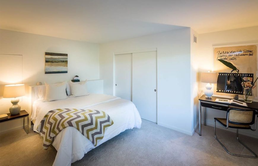 The two sections of this bedroom are separated by a swath of open space, but united by a similar color palette. This is an example of a dark bedroom. While the bedroom lacks recessed lighting or natural light, it can be made to seem lighter with white paint, bedding, and a few table lamps.