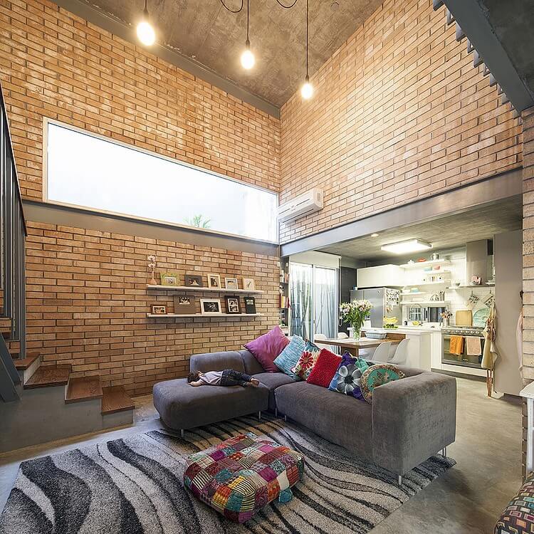 The central living room space reaches two stories high, with brick walls surrounding the metal structure on all walls. L-shaped sectional with variety of throw pillows stands next to multicolored ottoman, with kitchen and dining space in background. The central living room space reaches two stories high, with brick walls surrounding the metal structure on all walls. L-shaped sectional with variety of throw pillows stands next to multicolored ottoman, with kitchen and dining space in background.
