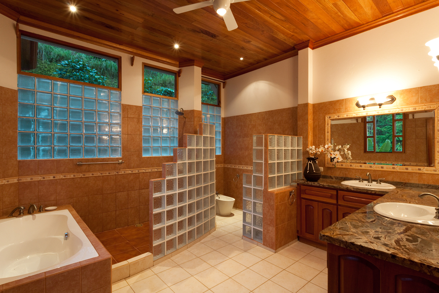 Intricate, lush bathroom stands artful brick cube walls between shower, toilet, and vanity spaces. Beneath a hardwood ceiling, dark wood cabinetry under dark marble countertops, light brown tiling, and white floors add variety of texture. Intricate, lush bathroom stands artful brick cube walls between shower, toilet, and vanity spaces. Beneath a hardwood ceiling, dark wood cabinetry under dark marble countertops, light brown tiling, and white floors add variety of texture.