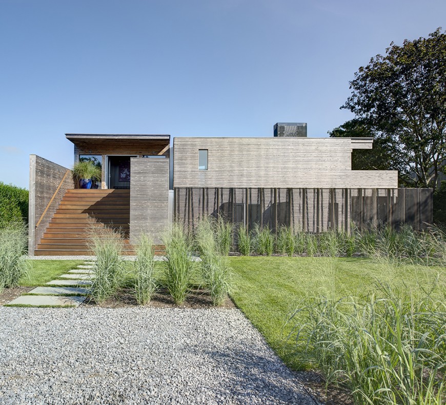 The home as seen approaching from the front. Stone path leads toward grand warm toned staircase between dividing walls of grey wood. The entire structure is minimal and angular, a flat presence hiding the complexity and ingenuity from this angle. The home as seen approaching from the front. Stone path leads toward grand warm toned staircase between dividing walls of grey wood. The entire structure is minimal and angular, a flat presence hiding the complexity and ingenuity from this angle.