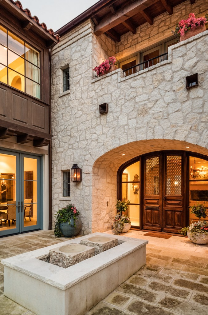 This broad, open entryway sets a double wide door beneath a stone arch, with partially obscured balcony overhead. Large stone bricks wrap a central fountain before the wood doors. This broad, open entryway sets a double wide door beneath a stone arch, with partially obscured balcony overhead. Large stone bricks wrap a central fountain before the wood doors.
