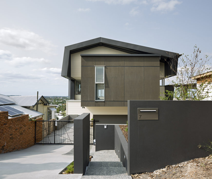 Front entryway view: we see the sloping drive and steps leading toward a concealed door. Above, the exterior in white and natural wood tones is wrapped in an angular grey roof. Front entryway view: we see the sloping drive and steps leading toward a concealed door. Above, the exterior in white and natural wood tones is wrapped in an angular grey roof.