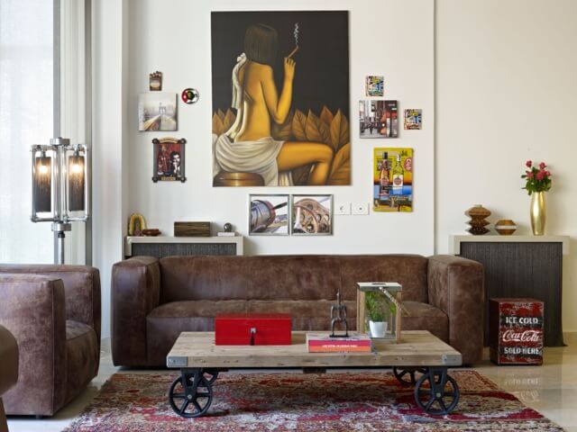 The living room area is dominated by contemporary brown leather sofa and matching armchair, around a natural wood table with black wheeled feet. Paintings hang on the wall next to one of several recycled industrial furniture pieces, a pipe-based lamp. The living room area is dominated by contemporary brown leather sofa and matching armchair, around a natural wood table with black wheeled feet. Paintings hang on the wall next to one of several recycled industrial furniture pieces, a pipe-based lamp.