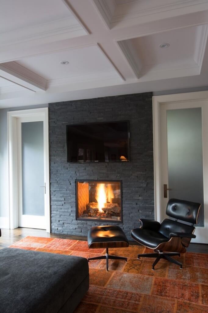 Two rooms are connected via this pass-through fireplace resting in a slate brick wall, flanked by fogged glass doors. A contemporary black leather, wood frame chair sits on a burnt orange rug beneath an elaborate, detailed ceiling design. Two rooms are connected via this pass-through fireplace resting in a slate brick wall, flanked by fogged glass doors. A contemporary black leather, wood frame chair sits on a burnt orange rug beneath an elaborate, detailed ceiling design.