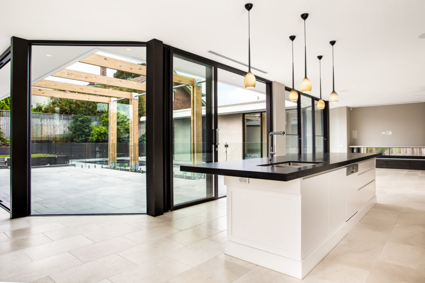 The wide open kitchen and living room space wraps around the new pool and patio area, seen through a bold swath of floor to ceiling glass panels. The large black countertop island seen at right defines the kitchen area. The wide open kitchen and living room space wraps around the new pool and patio area, seen through a bold swath of floor to ceiling glass panels. The large black countertop island seen at right defines the kitchen area.