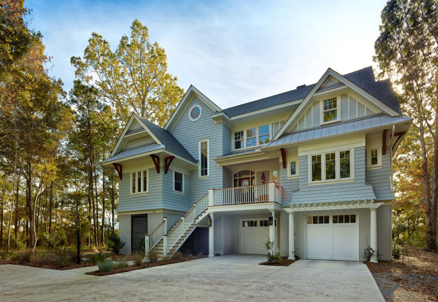 Standing brightly apart from its wooded surroundings, the tall home stands elevated above a concrete foundation with two garage stalls beneath the main body. Front entrance stands on a balcony wrapped in white and natural wood railings. Standing brightly apart from its wooded surroundings, the tall home stands elevated above a concrete foundation with two garage stalls beneath the main body. Front entrance stands on a balcony wrapped in white and natural wood railings.