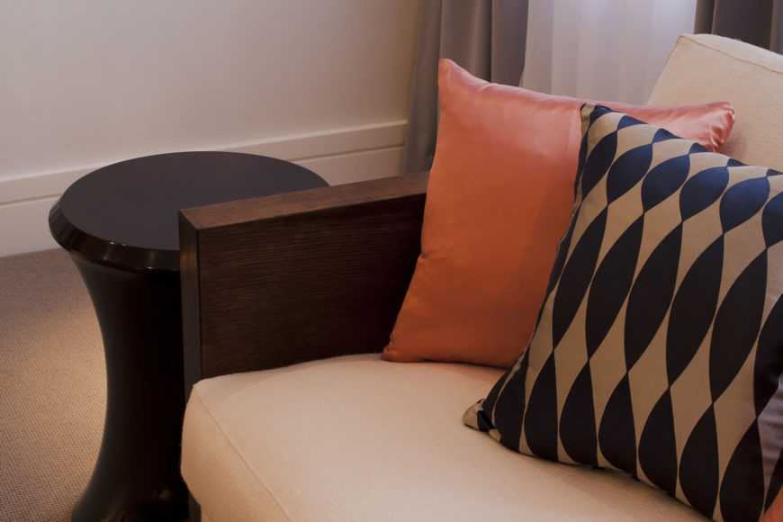 A close view of the sofa highlights the rich wood tones and contrast of patterned and colored pillows over the beige cushioning, which matches the carpet. A close view of the sofa highlights the rich wood tones and contrast of patterned and colored pillows over the beige cushioning, which matches the carpet.
