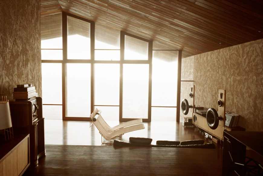 Looking down to the living room level, we see a pair of chaise lounges facing a bespoke wood and external speaker music setup, with sunlight glowing through the wood framed full height glass. Looking down to the living room level, we see a pair of chaise lounges facing a bespoke wood and external speaker music setup, with sunlight glowing through the wood framed full height glass.