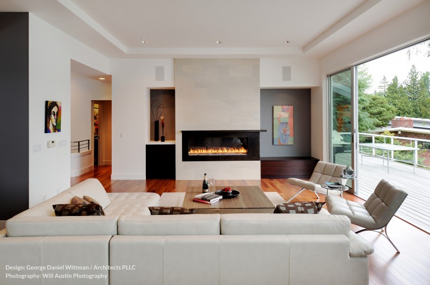 The living room opens almost entirely to the upper balcony, with large sliding glass panels as seen at right. A modern gas fireplace dominates the view, while white leather gracing the sectional and chairs contrasts with rich hardwood flooring. The living room opens almost entirely to the upper balcony, with large sliding glass panels as seen at right. A modern gas fireplace dominates the view, while white leather gracing the sectional and chairs contrasts with rich hardwood flooring.