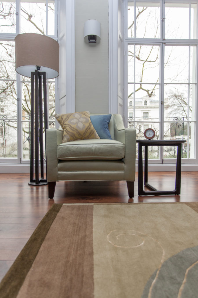 The living room holds earth toned seating and area rug over rich hardwood flooring. This club chair is flanked by an angular side table and floor lamp, with large, full height windows in background. The living room holds earth toned seating and area rug over rich hardwood flooring. This club chair is flanked by an angular side table and floor lamp, with large, full height windows in background.
