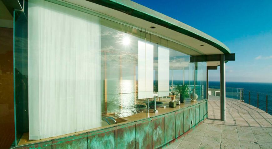On the upper level stone balcony wrapping the ocean side of the home, we see the gentle curve of the structure, sandwiching the large format glass panels between layers of exposed structure. On the upper level stone balcony wrapping the ocean side of the home, we see the gentle curve of the structure, sandwiching the large format glass panels between layers of exposed structure.