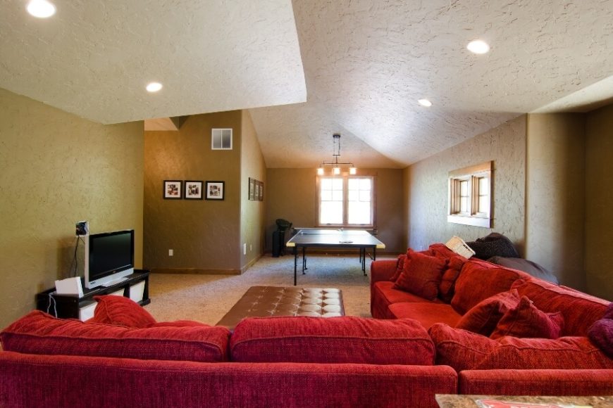 A view of the family room's bright red velour sectional from the opposite side of the room, showing the spacious game space that is occupied by a ping-pong table. The faux-leather ottoman is light enough to be easily moved out of the way if needed when playing Wii. A view of the family room's bright red velour sectional from the opposite side of the room, showing the spacious game space that is occupied by a ping-pong table. The faux-leather ottoman is light enough to be easily moved out of the way if needed when playing Wii.