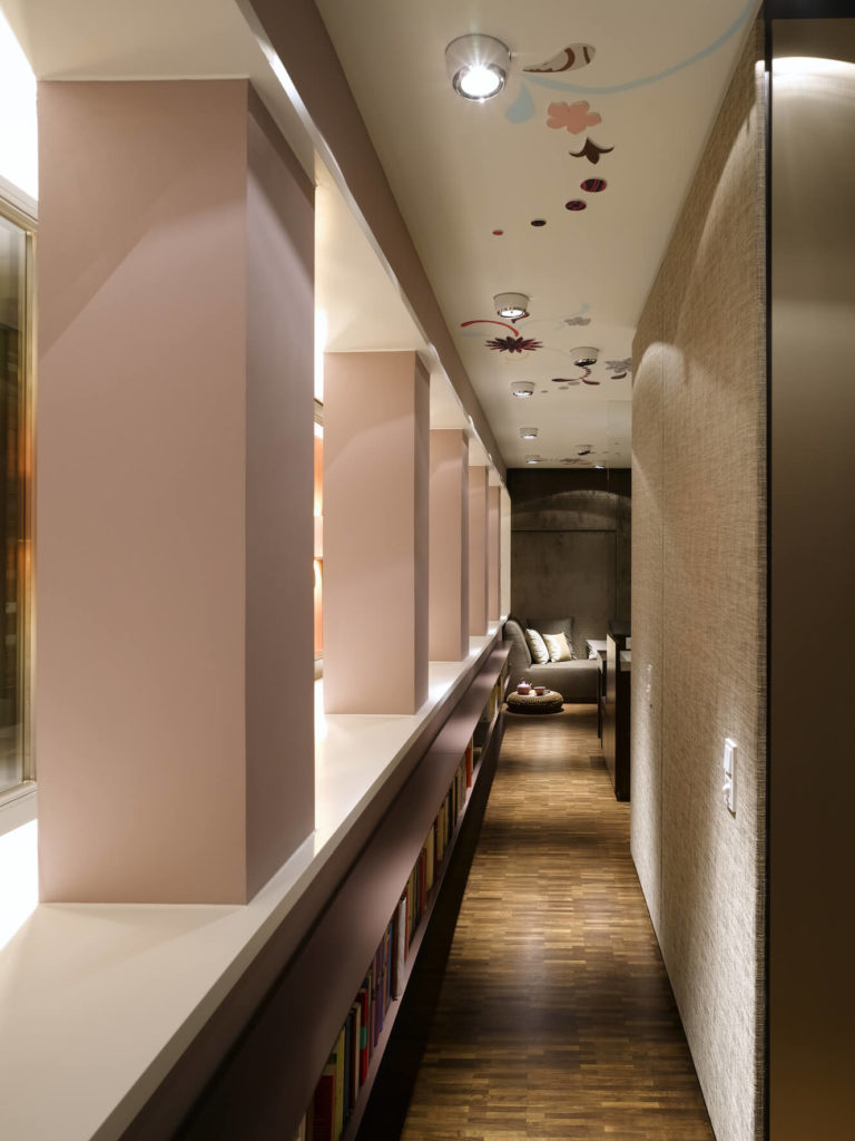 We begin in a hall lined with book shelving. This low wall divides the space, leading toward a living room area at the far end. We begin in a hall lined with book shelving. This low wall divides the space, leading toward a living room area at the far end.