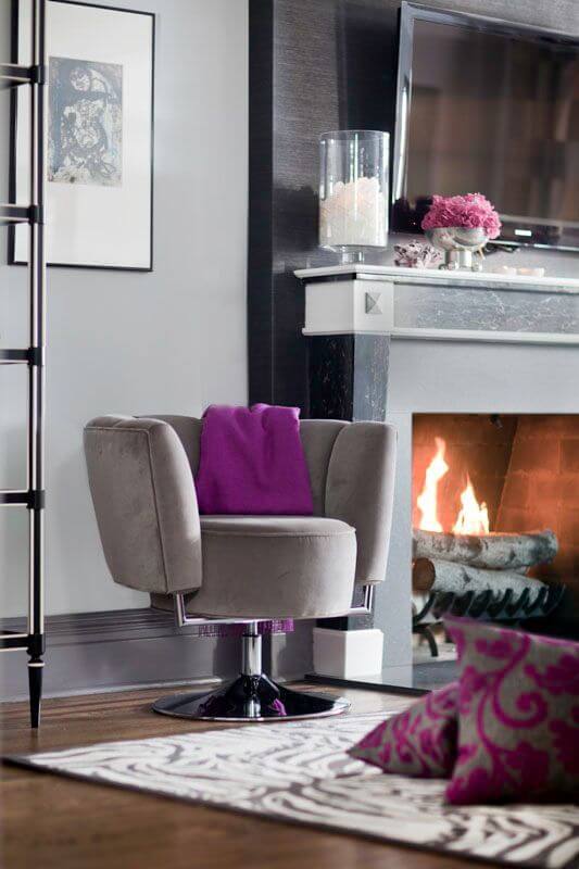 A close up on the living room's wood burning fireplace and the modern beige suede chair next to it. A fringed cashmere throw blanket in a beautiful bold berry purple is slung over the back of the rounded chair. The beautiful black soapstone around the fireplace adds an air of elegance. A close up on the living room's wood burning fireplace and the modern beige suede chair next to it. A fringed cashmere throw blanket in a beautiful bold berry purple is slung over the back of the rounded chair. The beautiful black soapstone around the fireplace adds an air of elegance.