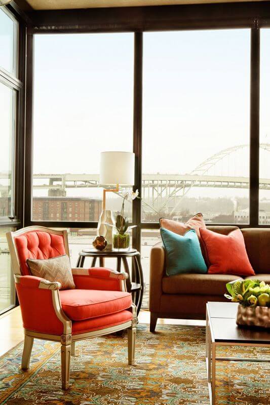 A close up of the living room's seating, showing the ornate armchair in a bright salmon with a beige accent pillow. The side table next to it is a barrel-shaped wooden table in a dark veneer with a modern white table lamp. A close up of the living room's seating, showing the ornate armchair in a bright salmon with a beige accent pillow. The side table next to it is a barrel-shaped wooden table in a dark veneer with a modern white table lamp.