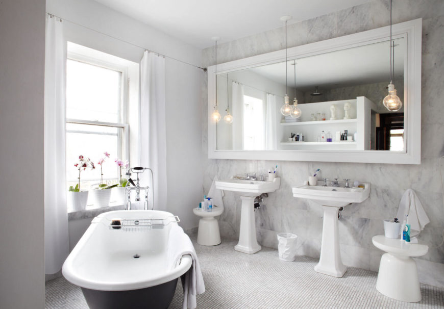 The bathroom houses an oversized mirror against a full height marble wall, with a pair of pedestal sinks in white below. The bathroom houses an oversized mirror against a full height marble wall, with a pair of pedestal sinks in white below.