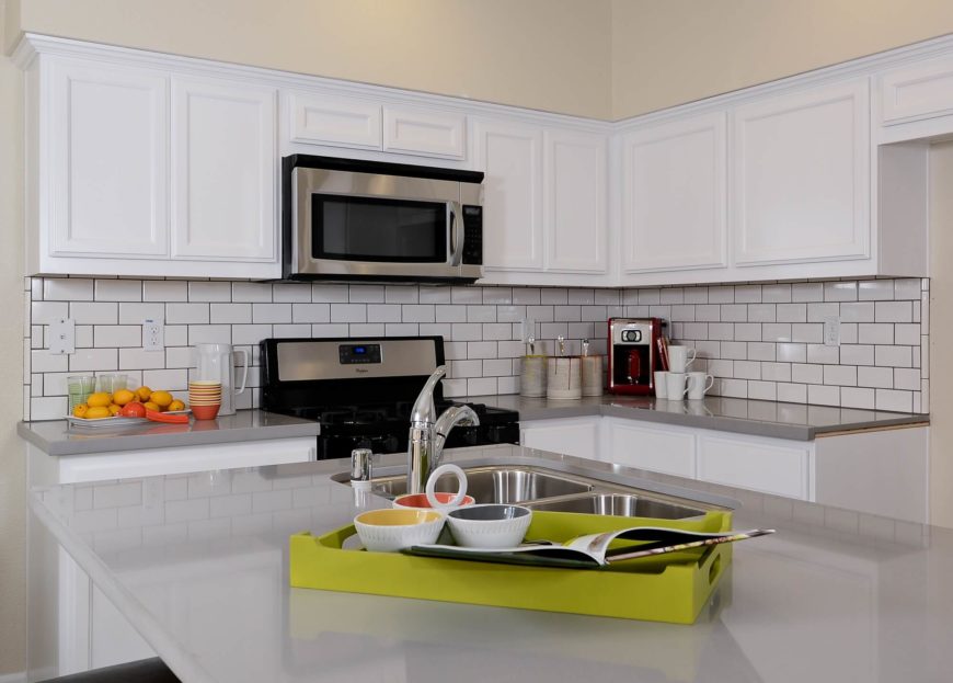The quartz countertops and white tile backsplash create contrasting texture within the white kitchen, sandwiched between layers of hardware-less cabinetry. The quartz countertops and white tile backsplash create contrasting texture within the white kitchen, sandwiched between layers of hardware-less cabinetry.