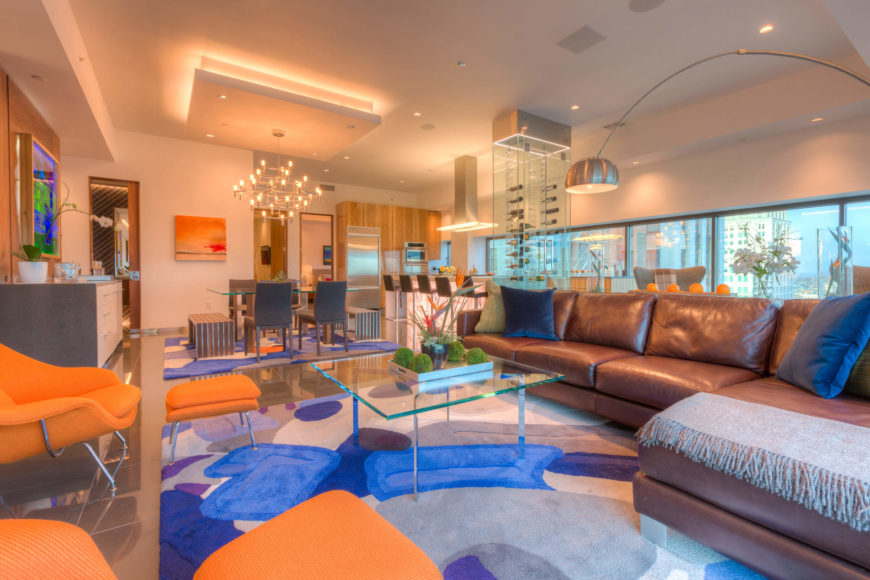 The living room within the open-plan space centers a leather sectional and bright orange lounges over a large multi-colored area rug, with the dining, kitchen, and full window wall in background. Exquisite details abound, including glass coffee table, large format tile flooring, and multiple layers of lighting throughout. The living room within the open-plan space centers a leather sectional and bright orange lounges over a large multi-colored area rug, with the dining, kitchen, and full window wall in background. Exquisite details abound, including glass coffee table, large format tile flooring, and multiple layers of lighting throughout.