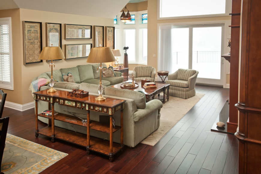 The living room centers on a large L-shaped sectional in light emerald tones paired with an armchair duo and large rich wood coffee table over a beige area rug. Deep, rich hardwood flooring fills the entire open-plan central space of the home, connecting the kitchen, through to the living room. The living room centers on a large L-shaped sectional in light emerald tones paired with an armchair duo and large rich wood coffee table over a beige area rug. Deep, rich hardwood flooring fills the entire open-plan central space of the home, connecting the kitchen, through to the living room.