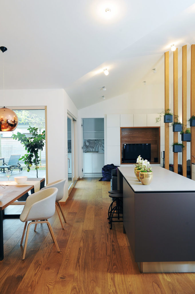 Viewing down the center of the large open-plan space, we see the bespoke grey kitchen island with white countertop standing across the rich hardwood floor from the light, natural wood dining table. The entire space glows with sunlight via expansive windows and sliding glass doors. Viewing down the center of the large open-plan space, we see the bespoke grey kitchen island with white countertop standing across the rich hardwood floor from the light, natural wood dining table. The entire space glows with sunlight via expansive windows and sliding glass doors.