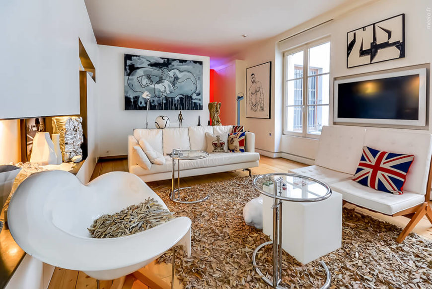 The living room furniture, in white and natural wood, centers on a recycled cloth rug over the hardwood flooring. To the left, interior lights glow over a carefully cut set of built-in shelving. The living room furniture, in white and natural wood, centers on a recycled cloth rug over the hardwood flooring. To the left, interior lights glow over a carefully cut set of built-in shelving.