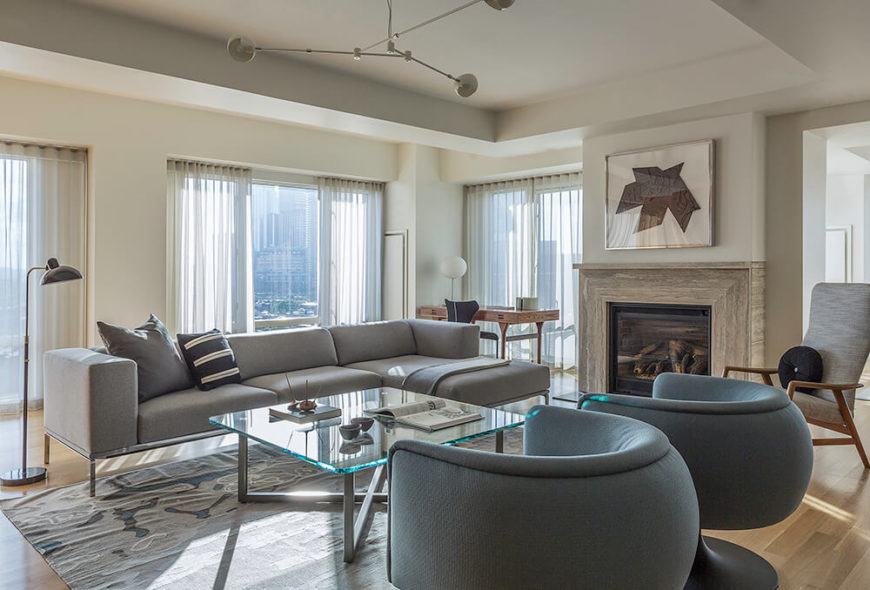 The living room centers on a sleek set of furniture, including a pair of spherical club chairs and a contemporary grey sectional sofa, plus a steel and glass coffee table at center. A marble fireplace stands between the doorway and one of several full height windows. The living room centers on a sleek set of furniture, including a pair of spherical club chairs and a contemporary grey sectional sofa, plus a steel and glass coffee table at center. A marble fireplace stands between the doorway and one of several full height windows.