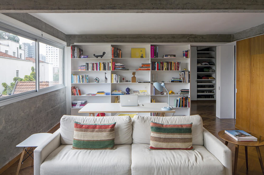 Beyond the living room sofa, we see a white minimalist writing desk with a full size built-in shelving unit at back. The shelving houses much of the color seen in the room, which takes the form of subtle accents and furniture. Beyond the living room sofa, we see a white minimalist writing desk with a full size built-in shelving unit at back. The shelving houses much of the color seen in the room, which takes the form of subtle accents and furniture.