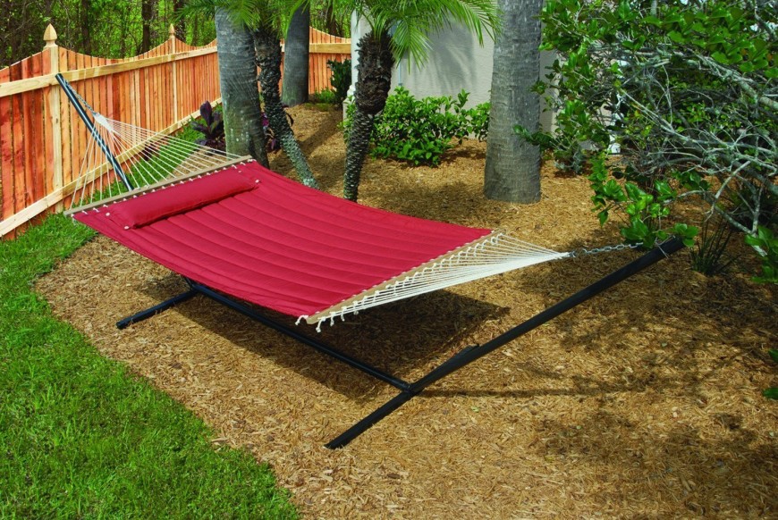 Another quilted hammock suspended on a freestanding metal frame. It's positioned over a mulched section of landscaping and shaded by several tall trees.