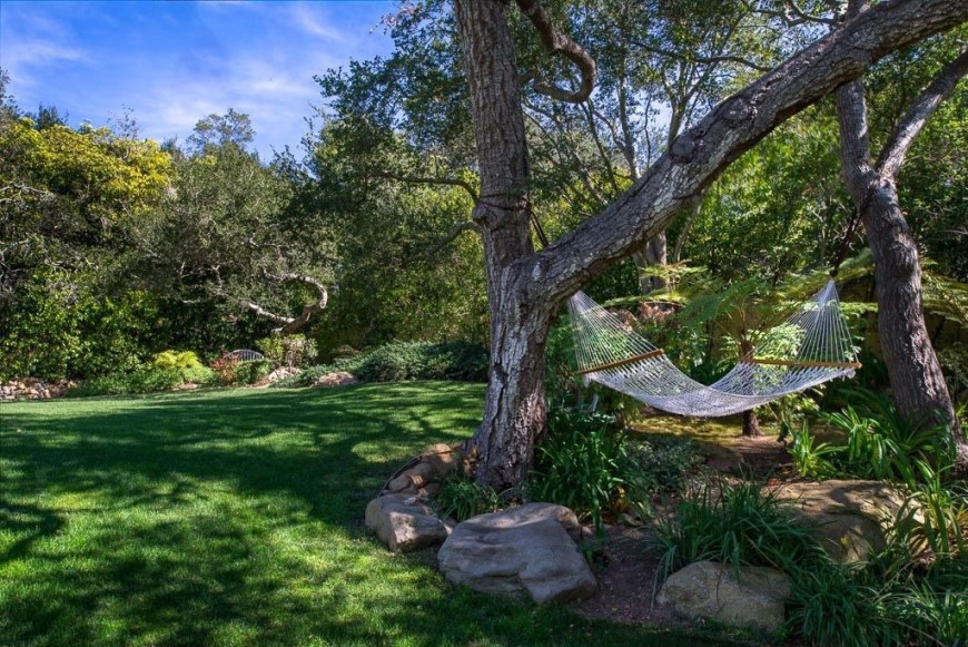 A simple, loose weave hammock hangs between two oak trees over a landscaped section of the yard looking out over the grass.