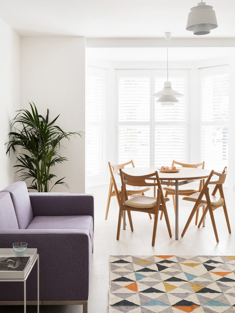 The dining area is part of the larger open plan space at the center of the home. Here we see how elements like the natural wood dining set, contemporary purple sofa, and multicolored rug offer texture and variety in the white space. The dining area is part of the larger open plan space at the center of the home. Here we see how elements like the natural wood dining set, contemporary purple sofa, and multicolored rug offer texture and variety in the white space.